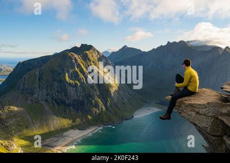 Mann, der am Rand eines Felsens sitzt und den Strand von Kvalvika in Lofoten, Norwegen, anschaut Stockfoto