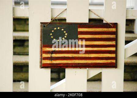 Am 4. Juli hängt eine koloniale amerikanische Flagge aus Farbe und Holz an einem weißen Zaun eines Hauses Stockfoto