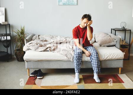 Junger Mann, der auf seinem Bett sitzt und morgens zu Hause aufwacht Stockfoto