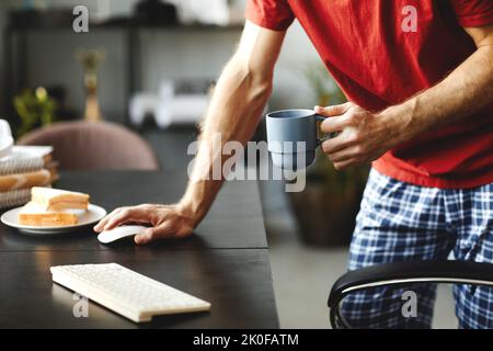 Nahaufnahme eines jungen Mannes, der morgens Kaffee trinkt und am Tisch einen Computer benutzt Stockfoto