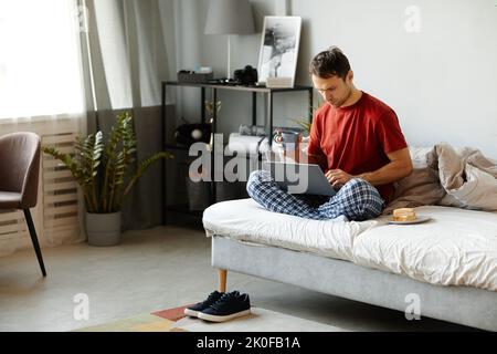 Der junge Mann benutzte einen Laptop und trank morgens in seinem Bett im Schlafzimmer Kaffee Stockfoto