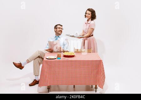 Porträt eines liebevollen Paares, eines Mannes, der Zeitung liest, einer Frau, die einen Tisch mit Kaffee und Kuchen serviert und das Frühstück auf weißem Hintergrund isoliert hat. Stockfoto