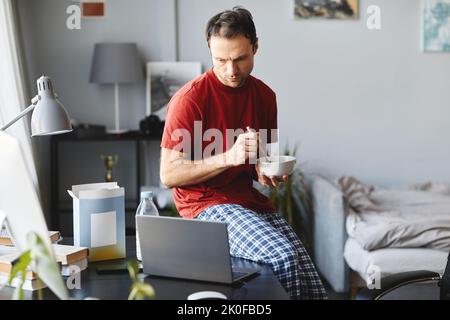 Der junge Mann, der Cornflakes zum Frühstück hatte, saß auf dem Tisch und schaute sich ein Video auf dem Laptop an Stockfoto
