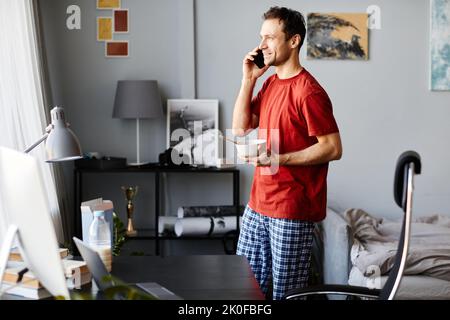 Junger Mann in Hauskleidung, der sich mit dem Handy unterhielt, während er im Zimmer frühstückte Stockfoto