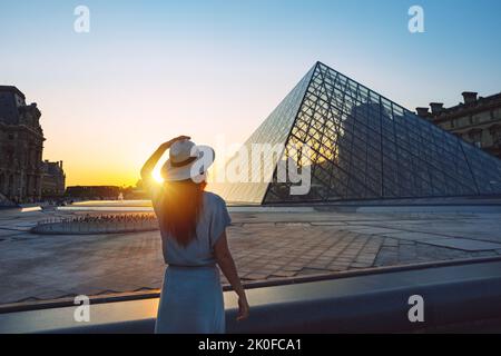 Junge Touristin in Sonnenhut, die bei Sonnenuntergang vor dem Louvre in Paris steht. Reisen in Frankreich, Tourismuskonzept Stockfoto