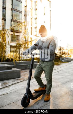 Junger Mann in warmer Kleidung verbindet Roller mit seinem Handy, um entlang der Straße zu fahren Stockfoto