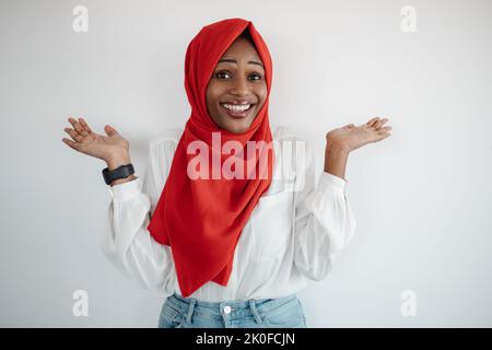 Studioaufnahme einer glücklichen schwarzen muslimischen Frau, die Handflächen ausbreitet, die die Hände hochhebt und die Kamera und die helle Studiowand anlächelt Stockfoto