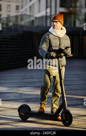 Junger Mann in warmer Kleidung mit Smartphone zu verbinden Roller, während auf der Straße stehen Stockfoto