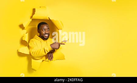 Glücklicher afroamerikanischer Mann, der durch ein Loch in gelbem Papier zur Seite schaut und mit dem Finger auf eine leere Stelle zeigt Stockfoto