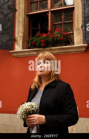 Unschärfe-Frau, die auf rotem Wandhintergrund steht und weiße Blumen hält. 40s seriöse Frau. Speicherplatz kopieren. Außen Porträt weiblich. Brautpaar. Blonde w Stockfoto