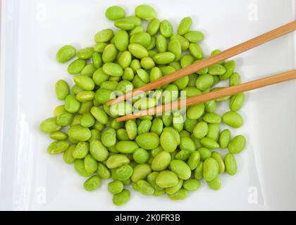 Draufsicht über Haufen geschälter Edamame-Bohnen auf einem weißen Porzellanteller mit darauf liegenden Stöcken. Pflanzliche Proteinquelle. Stockfoto