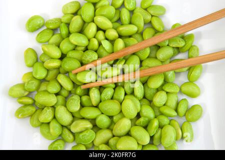 Draufsicht über Haufen geschälter Edamame-Bohnen auf einem weißen Porzellanteller mit darauf liegenden Stöcken. Pflanzliche Proteinquelle. Stockfoto