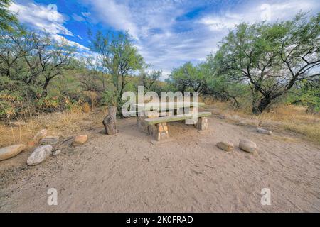 Picknicktisch auf einem sandigen Boden mit großen Steinen in Tucson, Arizona. Betontisch mit Sitzen in der Nähe des Grases mit Bäumen und saguaro Kaktus gegen Th Stockfoto