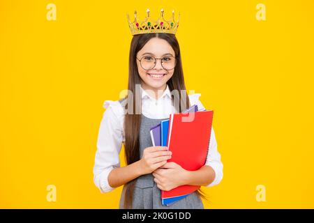 Schulmädchen in Schuluniform und Krone feiern den Sieg auf gelbem Hintergrund. Schulkind hält Bücher. Bildungsabschluss, Sieg und Erfolg. Stockfoto