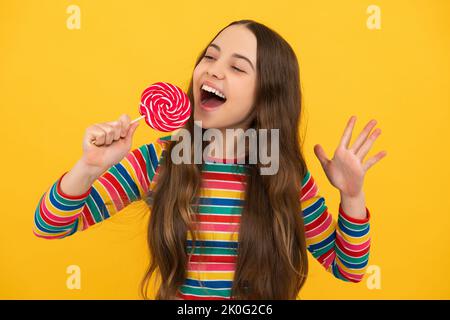 Lustiges Kind mit Lutscher auf gelbem isoliertem Hintergrund. Süßes Kindheitsleben. Teen Mädchen mit yummy Karamell Lollipop, Süßwarenladen. Aufgeregtes Gesicht Stockfoto