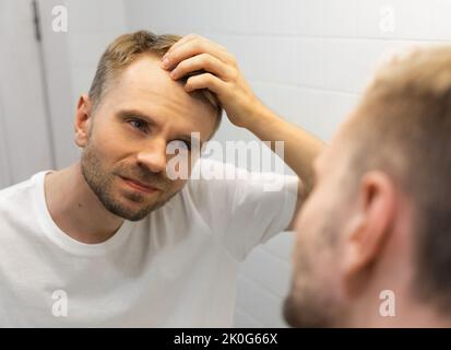 Weißer Mann mittleren Alters mit einem kurzen Bart sieht sich sein Haar im Spiegel im Badezimmer an und macht sich Sorgen wegen einer Glatze. Stockfoto
