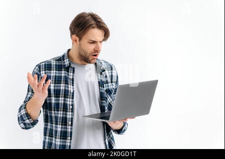 Frustriert verwirrt verwirrt verwirrt kaukasischen stilvollen jungen Mann, auf einem weißen isolierten Hintergrund stehend, hält einen offenen Laptop, überrascht schaut auf den Bildschirm, sehen unerwartete Nachrichten, Nachricht, gestikulierend mit der Hand Stockfoto