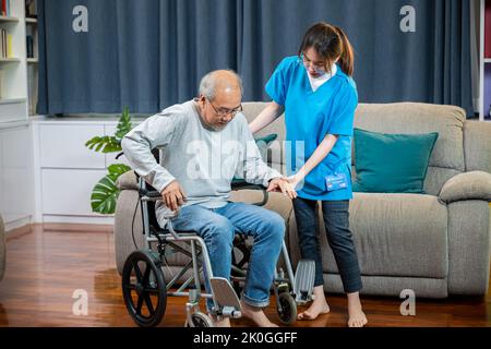 Asiatische Krankenschwester, die älteren Patienten hilft, aus dem Rollstuhl aufzustehen, um zu Hause zu gehen, lächelnder Arzt unterstützt den alten Mann beim Aufstehen, er Stockfoto