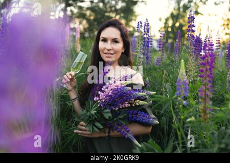 Eine attraktive Frau in einem Sommerfeld mit einem Glas Champagner und einem Bouquet von wilden Lupinus-Blumen. Vorderansicht. Stockfoto