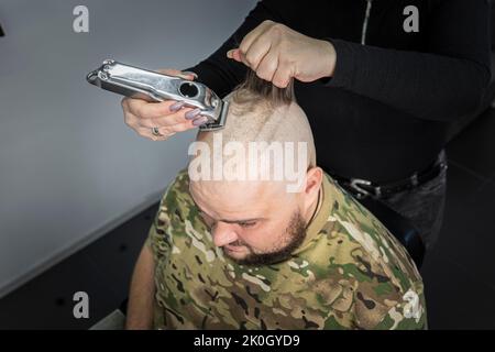Ein junger Mann in Militäruniform rasiert sich zum Militärdienst den Kopf kahl. Ein Mann mit Bart bekommt einen Haarschnitt in einem Friseurladen. Stockfoto