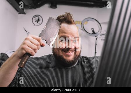 Bärtiger Hipster-Mann, der sich den Kopf rasieren ließ, wobei eine Axt zum Spiegel schaute. Ein strenger Mann rasiert sich den Kopf auf seinen Blick in den Spiegel. Stockfoto