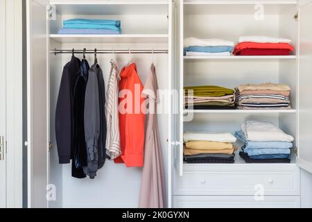 Regale mit farbenfroher Kleidung in einem weißen Kleiderschrank in Nahaufnahme, minimalistischer Stil Stockfoto