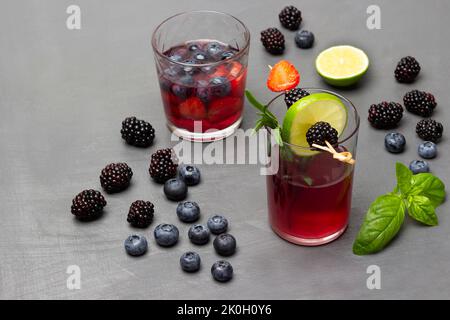 Brombeeren, Heidelbeeren, grüne Basilikumblätter auf dem Tisch. Zwei Gläser Beerengetränk. Brombeere und Limettenkeil am Spieß. Draufsicht. Grauer Hintergrund. Stockfoto