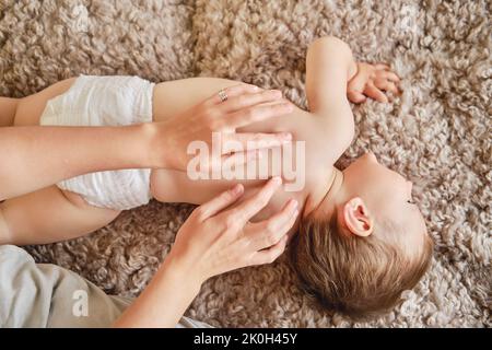 Frau Mutter tut Rückenmassage zu glücklichen Kleinkind Baby liegen auf beige Bettdecke. Mutter Masseur und lächelnd Kind Junge. Kind im Alter von einem Jahr und zwei Monaten Stockfoto
