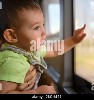 Kleinkind schaut aus dem Busfenster, während es in den Armen seiner Mutter sitzt. Kind im Transport mit einem Elternteil, Nahaufnahme Stockfoto