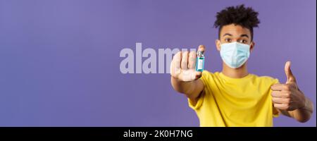 Nahaufnahme des aufrührenden jungen hispanischen Mannes in der medizinischen Maske, die Ampulle mit einem Kovid19-Impfstoff hält, Coronavirus-Medikament, zeigt Daumen hoch und wird angeschossen Stockfoto
