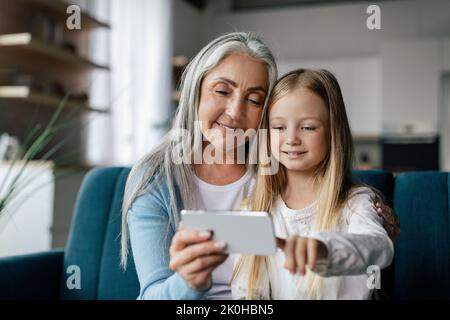 Fröhliche kaukasische kleine Enkelin und gealterte Oma sehen sich Videos an, rufen online am Telefon an, nutzen neue App Stockfoto