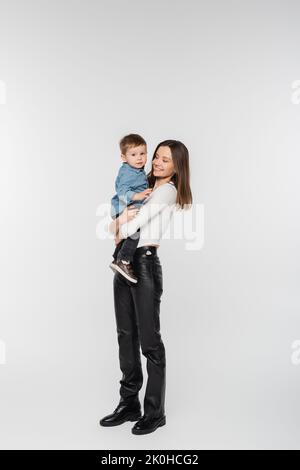 Volle Länge der jungen und glücklichen Frau hält Kleinkind Sohn auf grauem Hintergrund, Stockbild Stockfoto