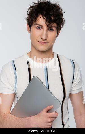 Trendy Brünette Mann in gestreiftem T-Shirt mit Laptop und Blick auf die Kamera isoliert auf grau, Stock Bild Stockfoto