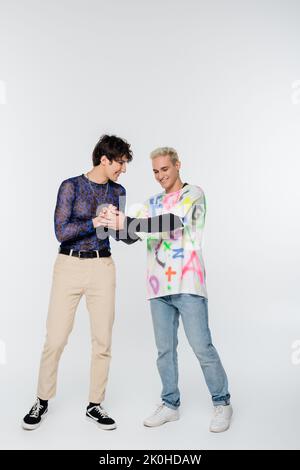 Volle Länge der positiven Homosexuell hält Hände von stilvollen queer Person auf grauem Hintergrund, Stockbild Stockfoto