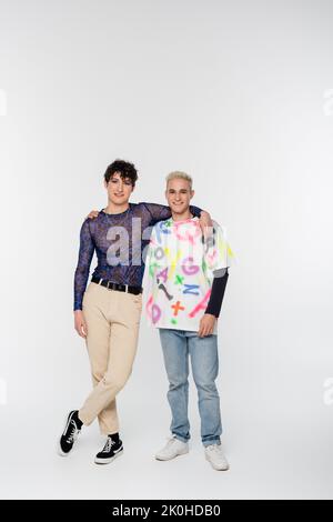 In voller Länge von jungen und fröhlichen queer Person mit Homosexuell Mann umarmt und Blick auf die Kamera auf grauem Hintergrund, Stockbild Stockfoto