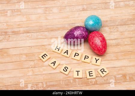 Drei helle Eier mit Happy Easter in Kacheln auf hellem Holzhintergrund geschrieben Stockfoto