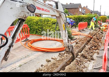 Minibagger mit rückwärtigem Schauspieler gräbt schmale Grabenrinne im Gehweg für orangefarbene Glasfaser-Breitbandkabel, die von den Arbeitern England UK positioniert wurden Stockfoto