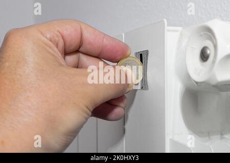 Nahaufnahme einer Männerhand, die eine Münze in den Schlitz des Heizkörpers im Zusammenhang mit der Preisenergiekrise in europa für eine teure Heizungs-cos einsteckt Stockfoto