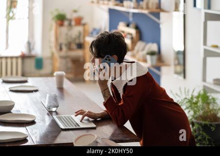 Junge weibliche Töpferunternehmerin, die ihren Laptop benutzt und telefoniert mit dem Kunden, um die Online-Bestellung zu überprüfen Stockfoto