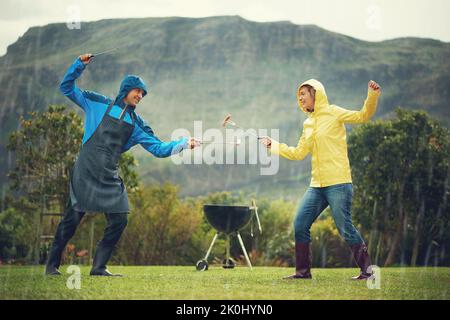 Ein fröhliches Paar, das im Regen grillt und mit Grillwerkzeugen fechten und Spaß haben. Stockfoto