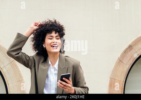 Glückliche Frau mit kabellosen Kopfhörern, die lachend Musik hört und mit ihrem Telefon tanzt, Konzept von Jugend und Technologie, Platz für Text kopieren Stockfoto