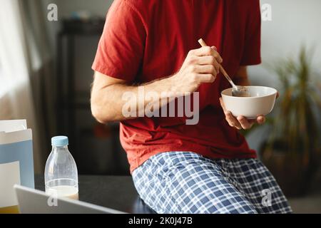 Nahaufnahme eines jungen Mannes, der eine Schüssel hält und Müsli zum Frühstück isst, während er sich ein Video auf dem Laptop ansieht Stockfoto