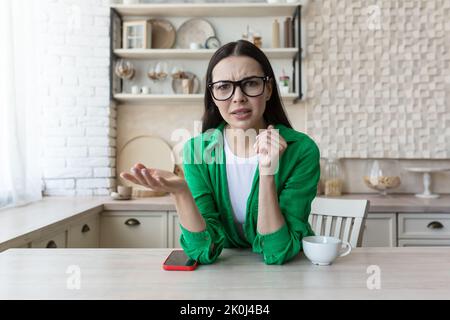 Bloggen. Webinar. Serious junge schöne Frau blogger im Gespräch mit der Kamera über Probleme. Verwirrt, rät. Sitzen zu Hause in der Küche am Tisch mit einem Telefon und einer Tasse Kaffee. Stockfoto