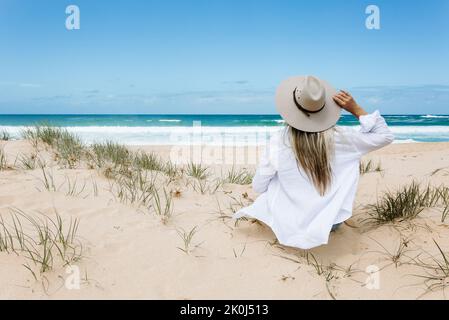 Sorglose Frau im Urlaub im fließenden weißen Hemd über Bikini-Schwimmkostüm, sitzt an einem sauberen Sandstrand mit blauem Himmel. Sie trägt einen Sonnenhut. Stockfoto