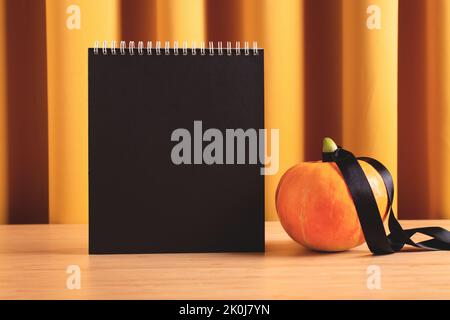 Halloween-Mockup mit leerer schwarzer Seite und Kürbis in dekorativer Wohneinrichtung. Traditionelle Urlaubssymbole und -Farben, Vorderansicht, Kopierbereich. Stockfoto