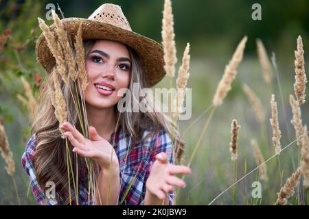 Schönes Cowgirl in Hut, kariertes Hemd und kurze Jeans bei Sonnenuntergang in einem ländlichen Feld Stockfoto