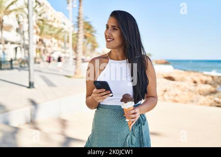 Junge hispanische Frau, die Smartphone benutzt und Eis am Meer isst Stockfoto