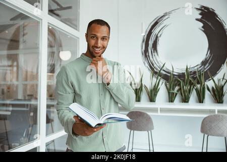 Lächelnder afrikanischer Geschäftsmann, der im modernen Büro steht und Buch hält Stockfoto