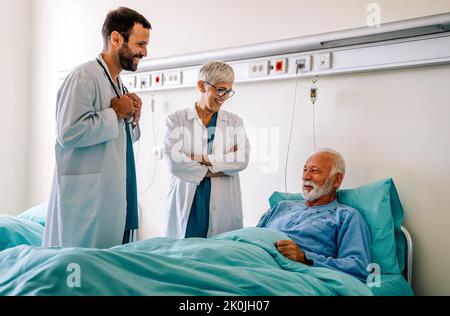 Arzt, der einen glücklichen älteren Patienten auf dem Krankenhausbett konsultiert. Konzept der Gesundheits- und Krankenversicherung Stockfoto