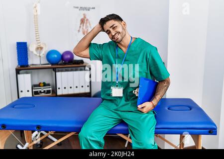 Der junge Physiotherapeut, der in der Schmerzklinik arbeitet, lächelt selbstbewusst und berührt die Haare mit der Hand nach oben, posiert attraktiv und modisch Stockfoto